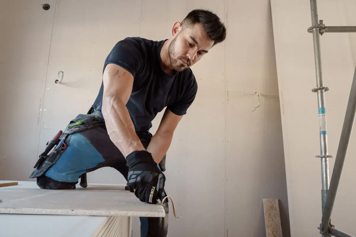 Drywall installer in work gloves scoring a gypsum panel with a utility knife on a cutting bench